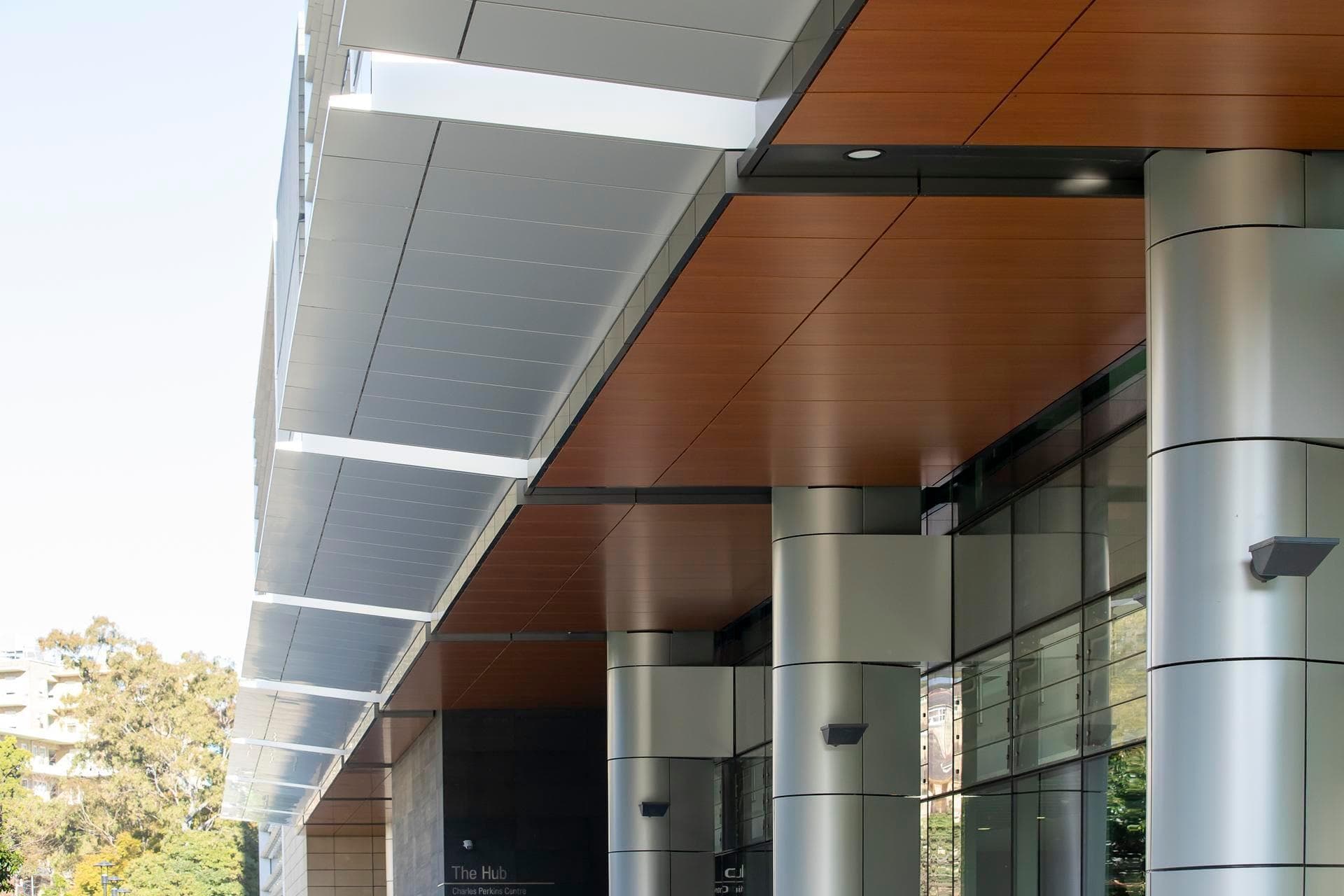 Exterior soffit of the Charles Perkins Centre featuring Alpolic NC cladding in silver and woodgrain panels supported by cylindrical columns.